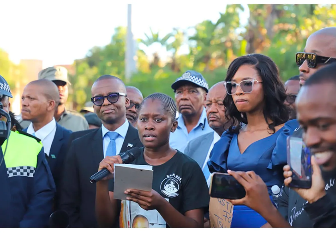 A young woman speaks into a microphone at a Re A Bua gathering, community members and officials standing with her